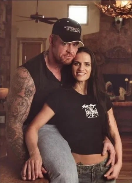 Mark Calaway in a black cap and black half sweat vest posing with his wife Sara Calaway in a black t-shirt and pants.